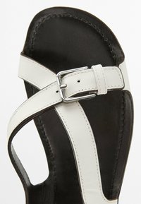 White leather sandal with a crisscross strap design, a silver buckle, and a black sole. Features smooth texture and precise stitching.
