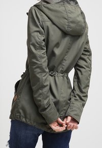 Olivově zelená bunda s kapucí vyrobená z hladkého materiálu. Má stahovaný pas, boční kapsy a značkový prvek poblíž lemu.