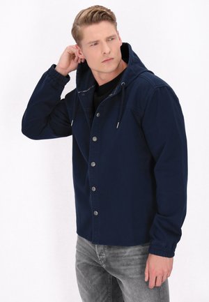 Jachetă cu kapușon de culoare albastru marin, cu butoane tip snap, manșete elastice și șnururi de ajustare. Textură de material neted și design cu o croială relaxată.