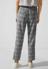 Persoon in grijs geruite rechte broek met elastieken tailleband en witte sneakers, staande tegen een effen lichte achtergrond.