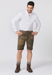 Braune Lederhosen mit strukturiertem Finish, Knopfverzierungen an den Seiten, kombiniert mit einem weißen Hemd und braunen Stiefeln.