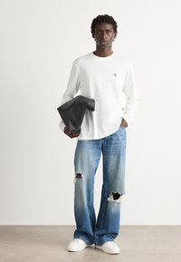Hvit langermet skjorte med logo, revne blå jeans og hvite joggesko; holder en sammenfoldet svart veske, står foran en nøytral bakgrunn.