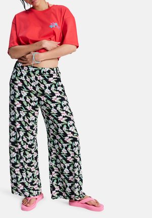 Nő piros rövidített pólóban, fekete, virágmintás, bő szárú nadrágban, rózsaszín platform szandálban és kék zsinórkarkötővel, karjai keresztben tartva áll.