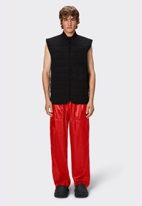 Gilet sans manches noir matelassé avec une fermeture éclair à l'avant et une poche, associé à un pantalon large rouge brillant et des chaussures noires texturées.