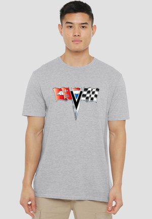 Junger Mann in einem grauen T-Shirt mit einem Design von drei Flaggen, darunter eine kariert aufgedruckte Rennflagge, auf der Brust vor einem einfarbigen Hintergrund.