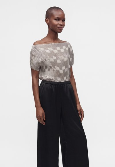 Femme portant un haut léger à motifs et épaules dénudées ainsi qu'un pantalon noir large, debout devant un fond clair uni.