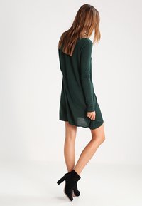 Grön stickad klänning med långa ärmar, en avslappnad passform och rund halsringning. I kombination med svarta ankelboots, visad från baksidan.
