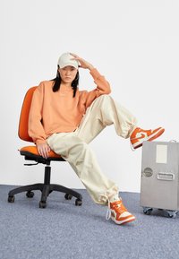 Orange sweatshirt, krämfärgade oversize-byxor och orange sneakers. Vit keps, hoops-örhängen, sittande i en svart kontorsstol, bredvid en grå väska.