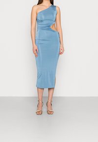 Robe bleue asymétrique avec découpe latérale, texture froncée et silhouette ajustée. Associée à des sandales métalliques à lanières.