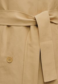 Μπεζ παλτό τρεντς με φαρδιά υφασμάτινη ζώνη δεμένη στη μέση και ορατό κουμπί σε λείο, ελαφρώς υφασμένο βαμβακερό ύφασμα.