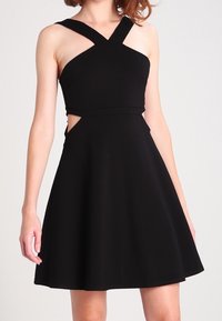 Svart klänning med en figursydd överdel och ett utsvängt kjoldel. Har korslagda band, utskärningar vid midjan och en slät, matt yta.