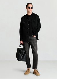Chemise noire boutonnée avec deux poches poitrine, t-shirt noir, jean gris, chaussures en daim marron et sac en cuir noir. Des lunettes de soleil complètent le look.