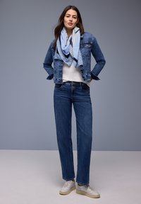 Femme portant une veste en jean, un jean bleu, des baskets blanches et une écharpe légère bleue à motifs, debout devant un fond gris uni.