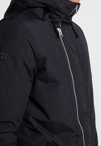 Hombre con una chaqueta negra con capucha, con una cremallera plateada en la parte delantera, tirador de cremallera y cordones negros visibles cerca del cuello.