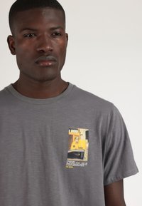 Camiseta gris de manga corta hecha de una tela suave, que presenta un pequeño gráfico de un coche vintage con acentos amarillos en la parte izquierda del pecho.