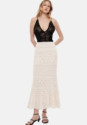 Donna che indossa un top senza maniche in pizzo nero con scollatura profonda e una lunga gonna crema all'uncinetto a vita alta e tacchi neri con cinturini.