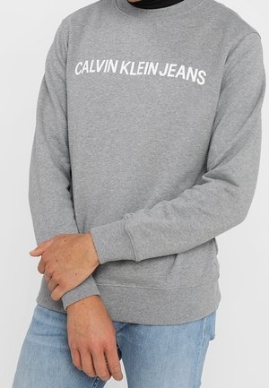 Osoba ubrana w szarą bluzę Calvin Klein Jeans z białym napisem logo i jasnoniebieskie dżinsy, z rękami skrzyżowanymi z przodu.