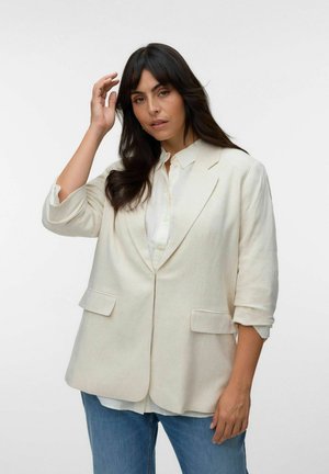 Femme aux longs cheveux foncés portant un blazer crème sur une chemise blanche et un jean bleu, posant avec une main près de la tête.