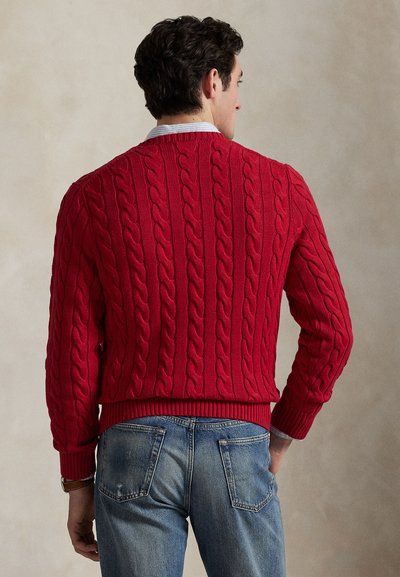 Uomo visto di spalle che indossa un maglione rosso a trecce sopra una camicia con colletto e jeans blu su uno sfondo beige uniforme.