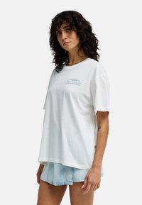 Mujer con cabello rizado y oscuro, vistiendo una camiseta blanca de Billabong y shorts de mezclilla claros, posando frente a un fondo blanco.