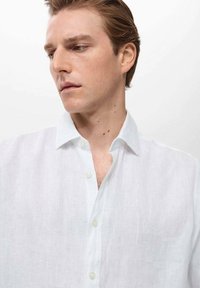 Camisa de lino blanca con un corte relajado, cierre de botones en la parte delantera y cuello clásico. La textura parece ligera con un brillo sutil.