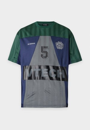 Ung mand der står iført en grøn og blå sportstrøje med nummer 5 og VTTECH-tekst, brune brede cargobukser og sorte sneakers.