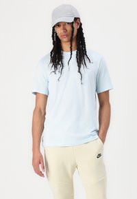Himmelblaues Baumwoll-T-Shirt mit kurzen Ärmeln, kombiniert mit beigen Jogginghosen, die ein schwarzes Nike-Logo tragen. Eine graue Mütze rundet das Outfit ab.