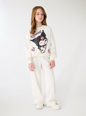 Ragazza giovane con felpa bianca con personaggio Kuromi e pantaloni bianchi a gamba larga, in piedi davanti a uno sfondo bianco uniforme.