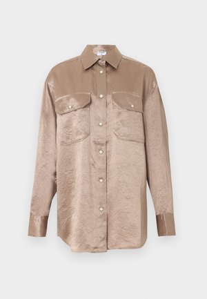 Model trägt ein glänzendes beiges Satinhemd und passende weite Hosen, kombiniert mit einem weißen T-Shirt und schwarzen spitzen Schuhen.