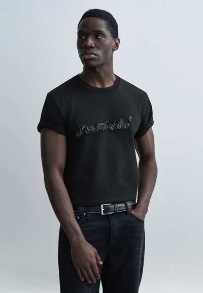 Μαύρο βαμβακερό T-shirt με γυρισμένα μανίκια και ανάγλυφο λευκό γραφικό κείμενο. Φορεμένο με σκουρόχρωμο τζιν και μαύρη ζώνη.