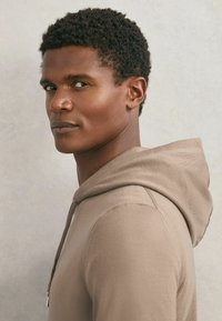 Ung mand med kort krøllet hår iført en beige hættetrøje, der kigger sidelæns med et neutralt udtryk mod en ensartet lys baggrund.