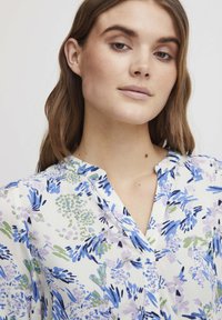 Blouse florale avec des motifs violets, bleus et verts sur fond blanc, dotée d'un décolleté en V et d'un tissu léger et doux.