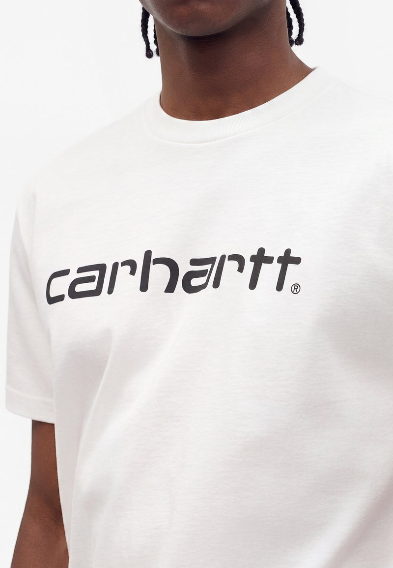 Person wearing a white t-shirt with short sleeves featuring a black curved Carhartt logo across the chest.