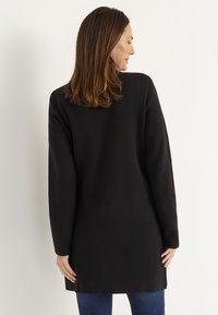 Svart långärmad tröja, texturerad tyg, rak passform, midjehöjd längd, sidofickor i fållen, bärs över jeans. Vy från baksidan.