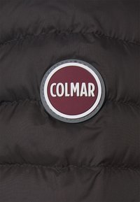 Svart pufferjacka med ett runt vinrött logopatch som säger "COLMAR" i vita bokstäver. Har en jämn textur och sydda paneler.