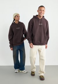 To modeller har på seg overdimensjonerte, mørkebrune hettegensere med lommer foran. De kombinerer dem med løse, blå jeans og kremfargede bukser, som fremhever varierte teksturer.