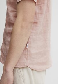Lichtroze linnen shirt met een afgeronde zoom, voorzien van een klein etiket aan de zijkant, en weergegeven op een neutrale achtergrond.