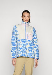 Sudadera de forro polar con una sección superior de crema acolchada, patrones florales azules y una cremallera rosa. Tiene un bolsillo en el pecho y un cuello alto.