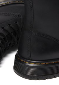 Botas de cuero negro con un acabado texturizado, con cordones redondos, ojales y una suela estriada con detalles de costura amarilla.