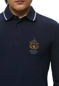 Polo a maniche lunghe color blu navy con righe bianche sul colletto, caratterizzato da un emblema ricamato con dettagli dorati e la scritta "Frecce Tricolori".