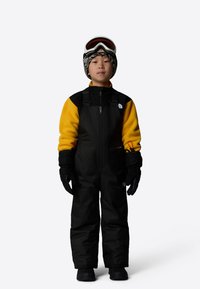 Black ski overall with a yellow fleece upper, featuring a front zipper, gloves, and ski goggles atop a patterned beanie.