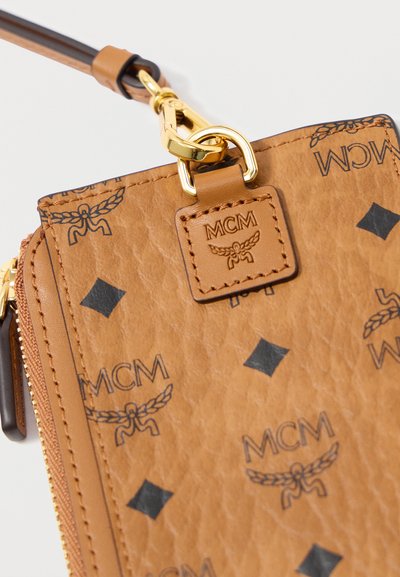 Καφέ δερμάτινο πορτοφόλι με υφή, που διαθέτει μαύρα λογότυπα MCM και διαμαντένια σχέδια, με χρυσό κούμπωμα και δερμάτινη ετικέτα.