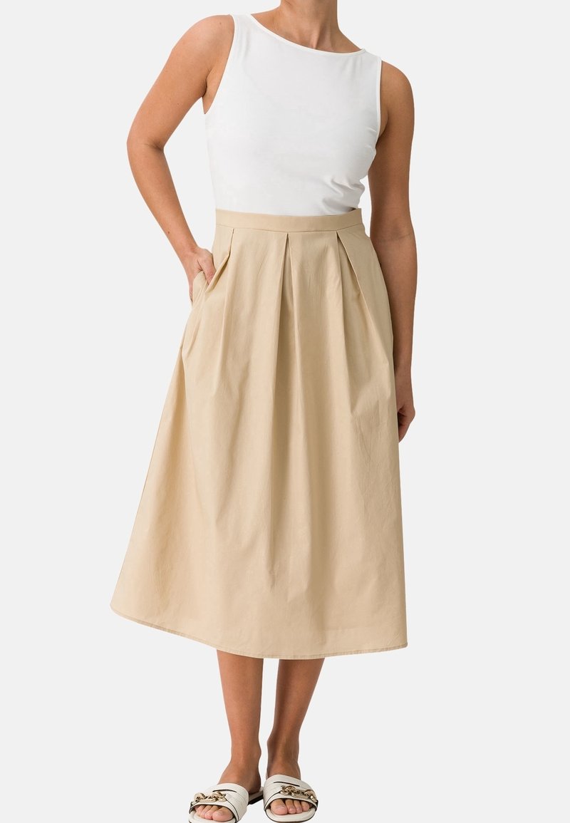 Femme portant un haut blanc sans manches, une jupe midi plissée beige avec poches, et des sandales blanches à enfiler ornées d'un détail chaîne en or.