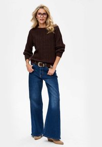 Brązowy sweter dzianinowy z teksturowym wzorem, połączony z niebieskimi szerokimi spodniami o wysokim stanie i tanimi botkami na obcasie. Noszone duże fioletowe okulary.