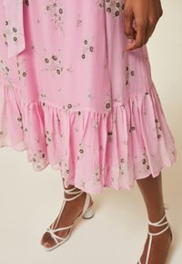 Rosa Blumenkleid mit Rüschensaum, verziert mit kleinen weißen und braunen Blumenmustern. Getragen zu weißen, schlichten Riemchen-High Heels.