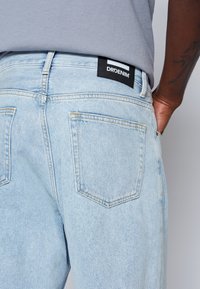 Ljusblå denimjeans med en svart DRDENIM-märke på midjebandet, med två bakfickor och en subtil blekt textur.