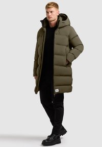 Abrigo puffer verde con capucha, que presenta un diseño acolchado, cierre de cremallera y bolsillos laterales. Lleva puesto sobre una blusa negra y pantalones negros.