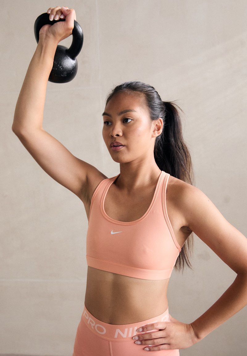 Black kettlebell raised above the head, worn by a person in a light peach sports bra and matching high-waisted leggings with white branding.