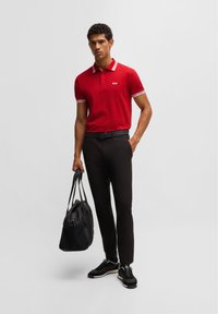 Rotes Poloshirt mit weißem gestreiftem Kragen und Bündchen, schwarze Hose und schwarze Sneakers. Die Person trägt eine schwarze Sporttasche.