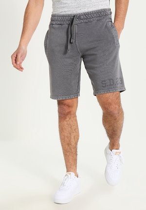 Mężczyzna w szarych szortach elastycznych w pasie z troczkiem i białych sportowych butach, idący na tle jednolitego tła.